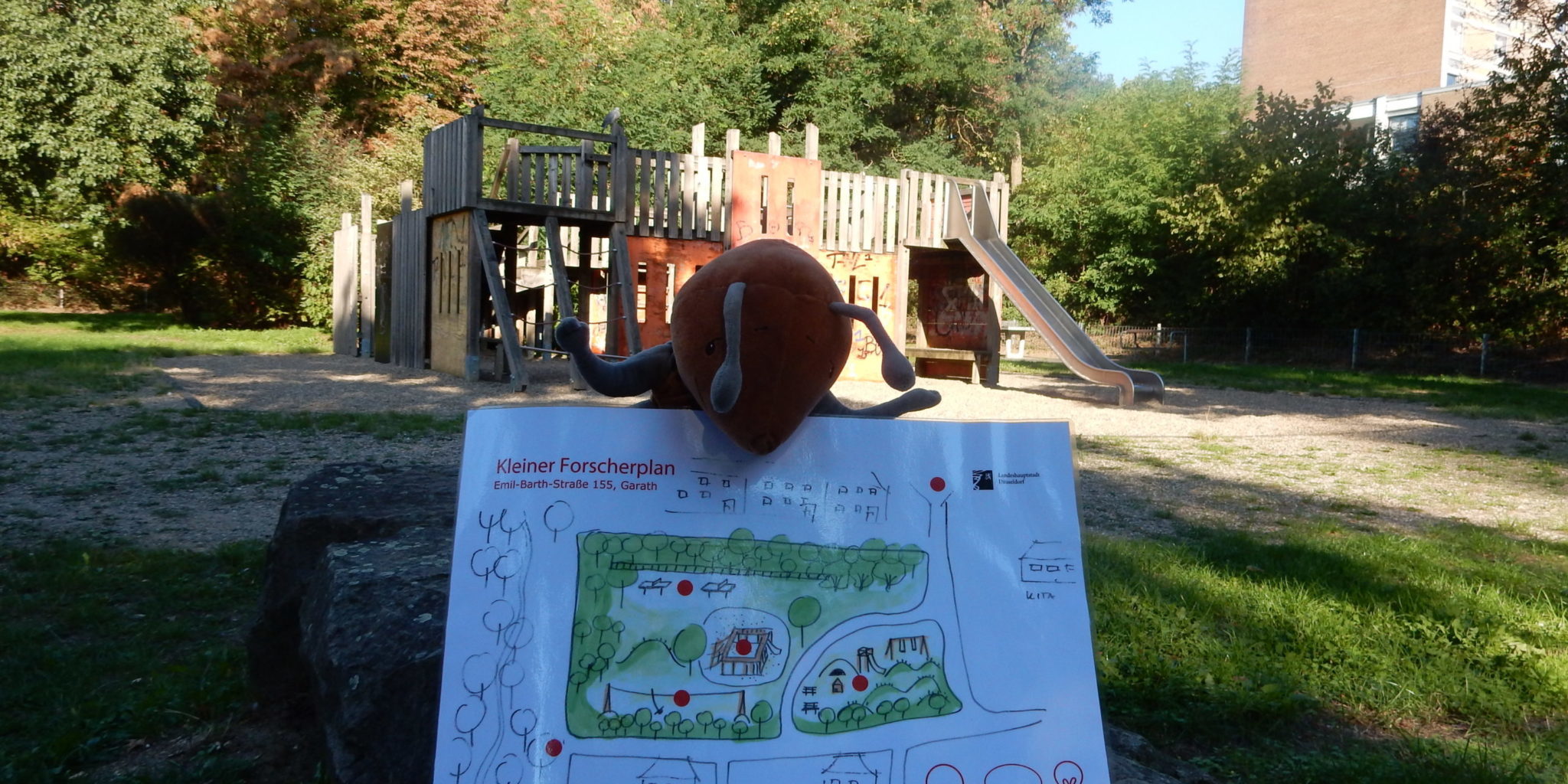 Plüschameise hält Plan vom Ameisenspielplatz vor der Kulisse des alten Spielplatzes