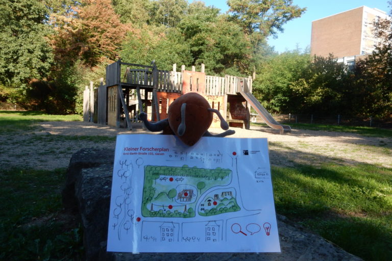 Plüschameise hält Plan vom Ameisenspielplatz vor der Kulisse des alten Spielplatzes