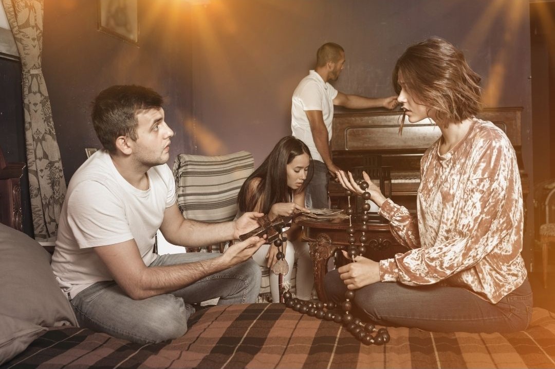 Das Bild zeigt eine Situation in einem Escape-Room. Der Raum ist dunkle u. Im Vordergrund sitzen sich rechts ein junger Mann und links eine junge Frau gegenüber auf dem Boden. Sie halten Perlenketten in der Hand. Ein Stück hinter den beiden sitzt mittig eine weitere junge Frau auf einen niedrigen Sessel. Sie betrachtet Zettel und Bücher auf einem niedrigen Tisch. Hinter ihr steht ein junger Mann vor einem Klavier.