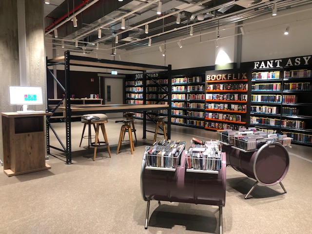 Bücherregal in einem großen Raum mit hohen Denken. In der Mitte steht ein hoher Tisch mit Hockern und vorne sind Kisten mit Büchern.