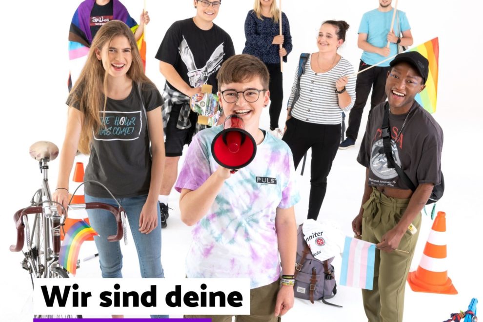 Vor weißem Hintergrund: Eine nachgestellte Straßendemonstration. Acht junge Menschen gucken lachend in die Kamera. Eine Teilnehmerin hält ein Megaphon, andere halten Regenbogenfahnen, ein Transparent "Kinder und Jugendrechte ins Grundgesetz" und eine Pappe mit "There is no planet B". Der Schriftzug über dem Motiv: "Wir sind deine Stimme!".