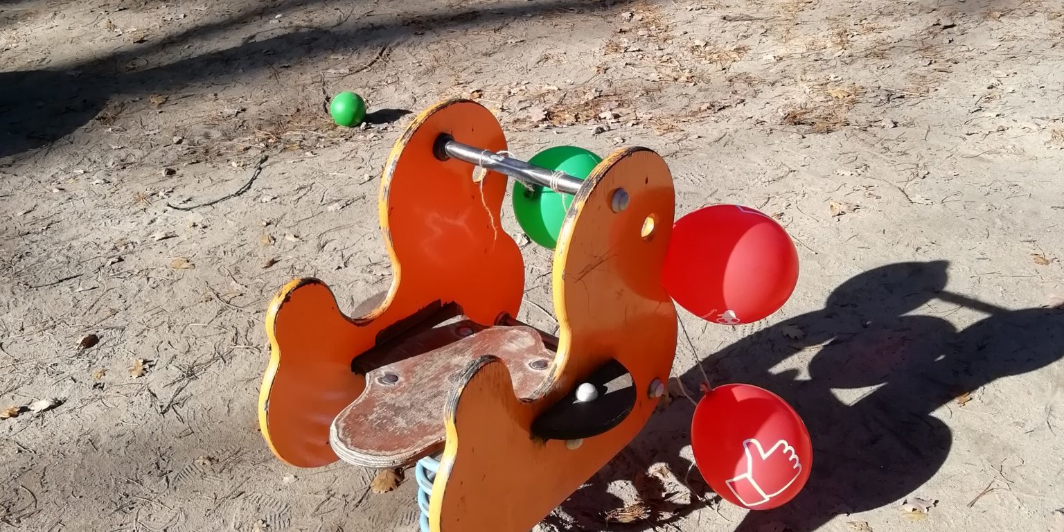 Ein gelb-rotes Wipptier an dem zwei rote und ein grüner Luftballon befestigt sind. Im Hintergrund sieht man einen Baum, eine Rutsche und zwei Schaukeln. Die Sonne scheint.