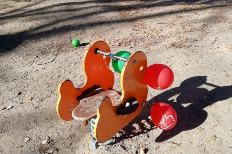 Ein gelb-rotes Wipptier an dem zwei rote und ein grüner Luftballon befestigt sind. Im Hintergrund sieht man einen Baum, eine Rutsche und zwei Schaukeln. Die Sonne scheint.