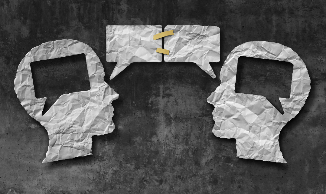 Auf einem schwarzen Hintergrund liegen aus Papier geschnittene Umrisse von Köpfen. DIe beiden Köpfe sind im Profit einander zugewendet. Zwischen beiden sind jeweils eine Sprechblase. Und auch im oberen Teil ihres Kopfes ist eine Sprechblase ausgeschnitten.