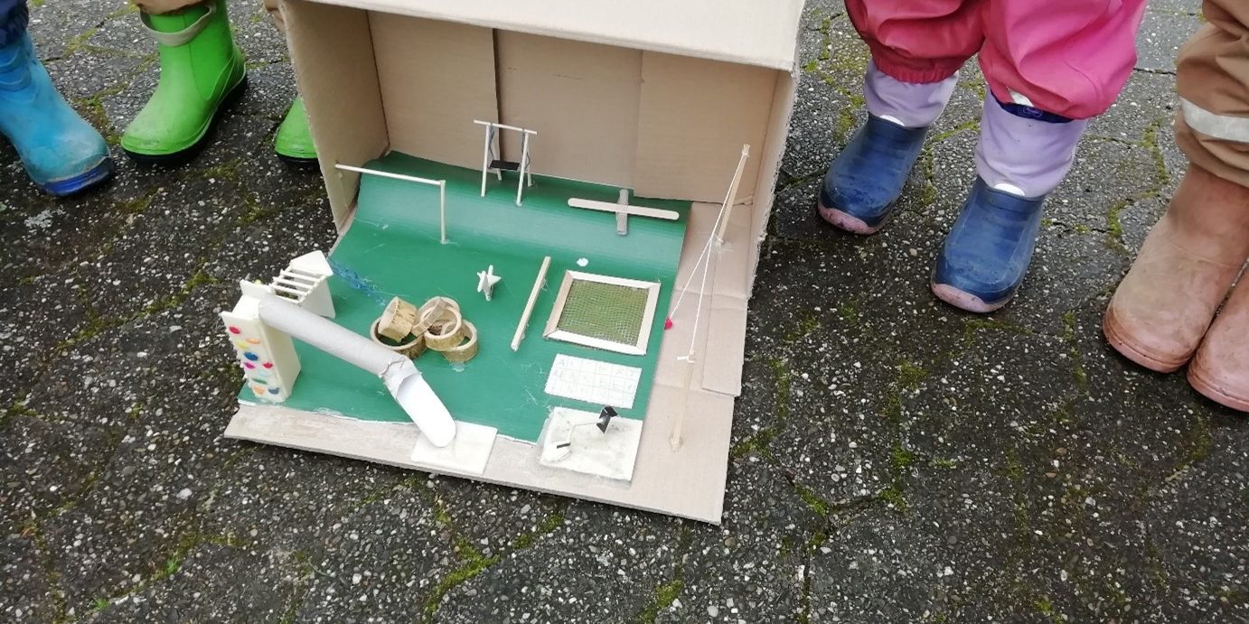 Kinder in Matschhosen und Gummistiefel stehen um ein sehr Detailgetreues Modell eines Spielplatzes herum.