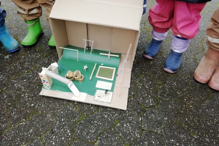 Kinder in Matschhosen und Gummistiefel stehen um ein sehr Detailgetreues Modell eines Spielplatzes herum.