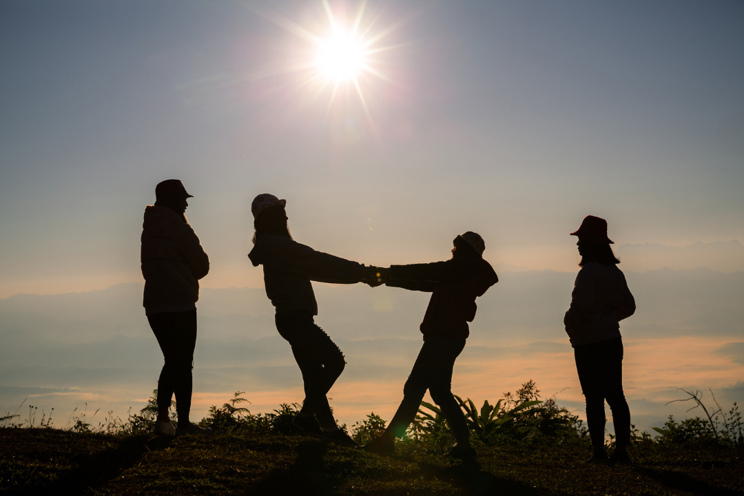 Vor einer niedrig stehenden Sonne sieht man 4 Personen, die im Gegenlicht nur als Umrisse erkennbar sind. Die beiden Personen in der Mitte halten sich an den Händen, sind einander zugewandt und lehnen sich zurück. Die anderen beiden stehen rechts und links daneben.