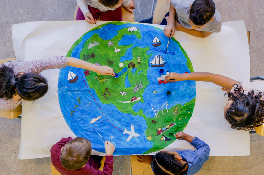 Fotoaufnahme von oben auf einen Tisch. Auf dem Tisch liegt ein großes, weißes Stoffstück auf dem der Planet Erde gemalt ist. Um den Tisch sitzen 6 Kinder, die die Erde malen.