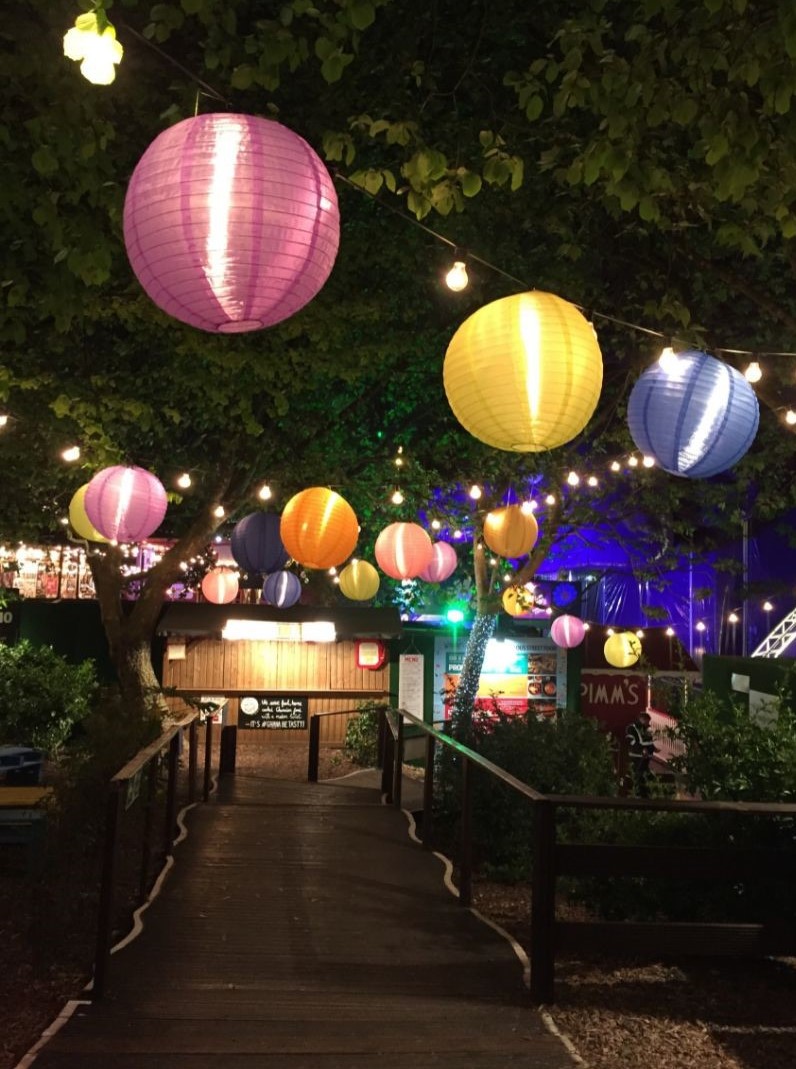 Bei Nacht sieht man einen Holzweg der zu einer Gartenhütte aus Holz führt. Der Weg ist mit bunten Lampen geschmückt.