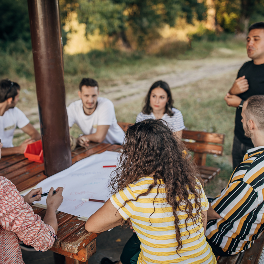 Eine Gruppe junger Menschen sitzt draußen an einem runden Holztisch. Eine Person rechts im Bild steht.