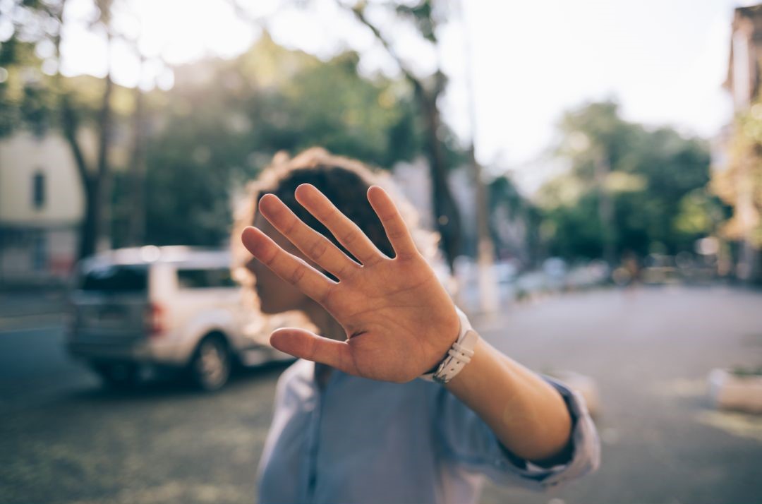 Eine junge Person steht auf einer Straße. Man sieht sie vom Oberkörper aufwärts. Sie hält ihre Handfläche ausgestreckt in Richtung Kamera und verdeckt damit ihr Gesicht.