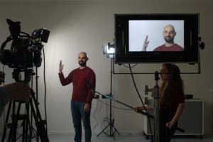 Im Vordergrund ist eine Kamera und ein Bildschirm, der das Bild der Kamera zeigt. Im Hintergrund steht mittag ein junger Mann, der in die Kamera spricht. Rechts an der Seite steht eine junge Frau, die ein langes Mikrophone hält.