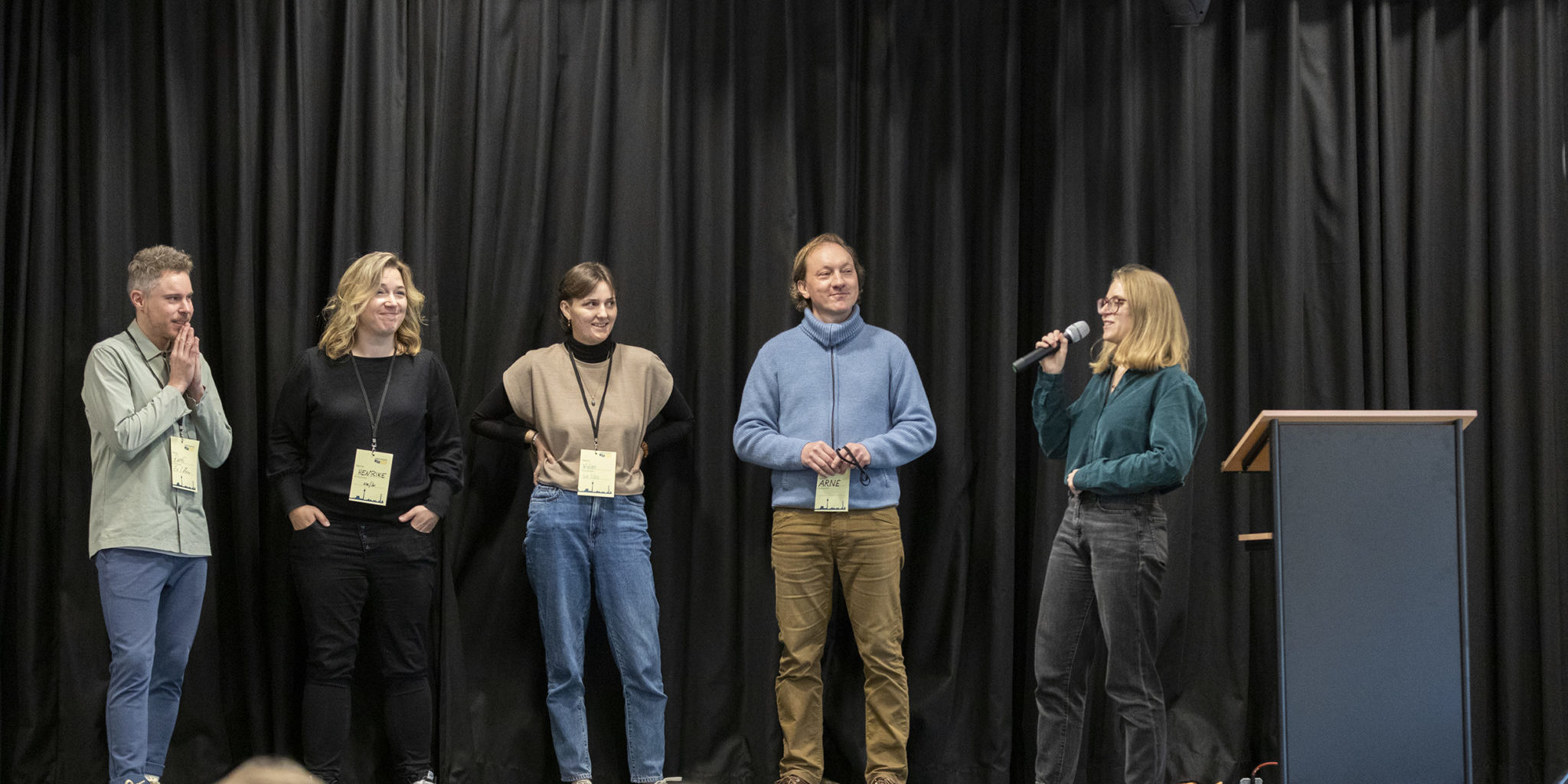 Auf einer Bühne stehen 5 Personen. Die Person ganz rechts hält ein Mikrophon.