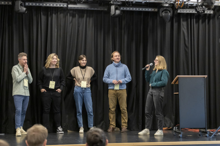 Auf einer Bühne stehen 5 Personen. Die Person ganz rechts hält ein Mikrophon.