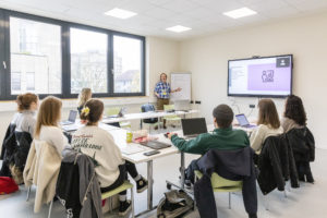 Blick in einen Seminarraum. Junge Menschen sitzen in einem U an Tischen. Vorne steht ein Mann neben einem Bildschirm und erklärt etwas.