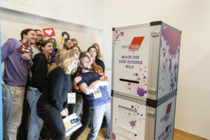 Eine Gruppe junger Menschen steht vor einer Fotobox und macht ein Gruppenbild.