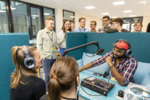 Drei jnge Menschen sitzen an einem Tisch auf dem Podcast-Equipment aufgebaut ist. der Tisch und die Sitze sind von 1,4m hochen Wänden umgeben. Andere Personen stehen um die Wände herum und schauen auf die drei Personen, die einen Podcast aufnehmen.