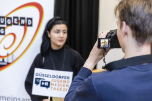Blick über die Schulter eines jungen Mannes, der eine Kamera auf eine Junge Frau richtet. Sie blickt in die Kamera und hält ein Schild auf dem Düsseldofer Jugendmedientage steht.