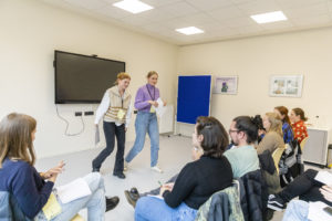 Zwei junge Frauen stehen von einer Gruppe sitzender Menschen und performen einen Text.