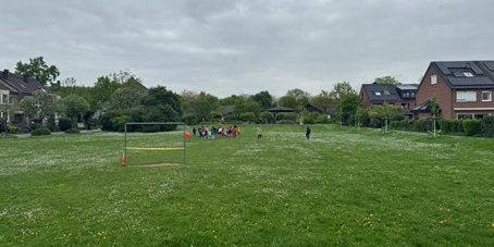 Auf der Wiese des Quartierplatzes ist in einiger Entfernung eine größere Grppe von Kindern und Erwachsenen zu sehen. Der Platz ist umgeben von Wohnhäusern und auf der Wiese steht ein Tor.